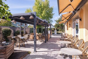 Un patio al aire libre con mesas y sillas, una pérgola con luces colgantes, y un edificio de fachada amarilla.