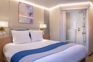 Habitación de hotel con una cama grande, dos lámparas de noche, un cuadro en la pared y zona de ducha.