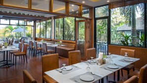 A restaurant with large windows, wooden furniture, and a view of tropical plants.