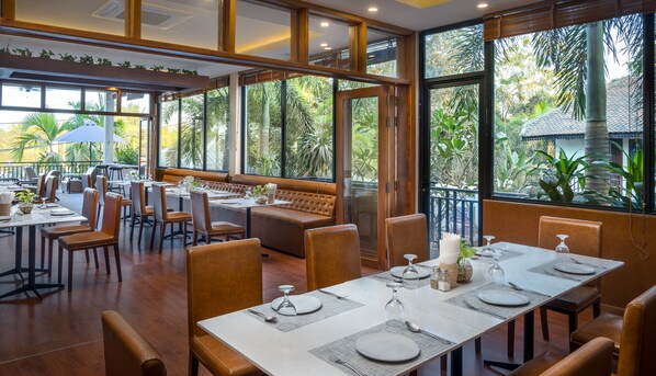 A restaurant with large windows, wooden furniture, and a view of tropical plants. - Amatak Boutique Hotel  (Siem Reap)