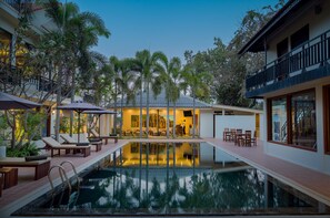 Un área junto a la piscina con sillones, comedor y un edificio con grandes ventanales.