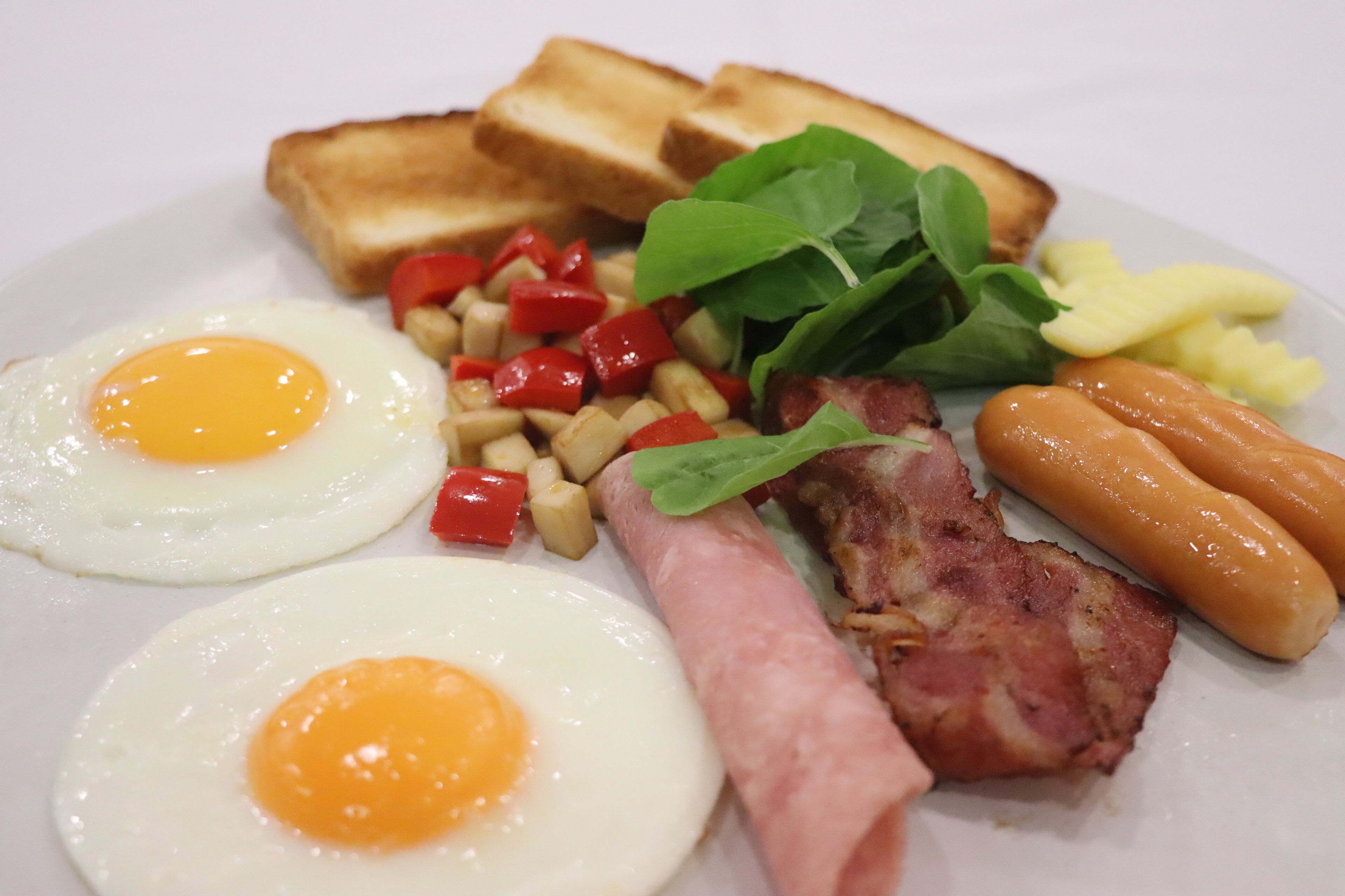 Un plato con dos huevos fritos, tostadas, jamón, salchichas y una guarnición de verduras.