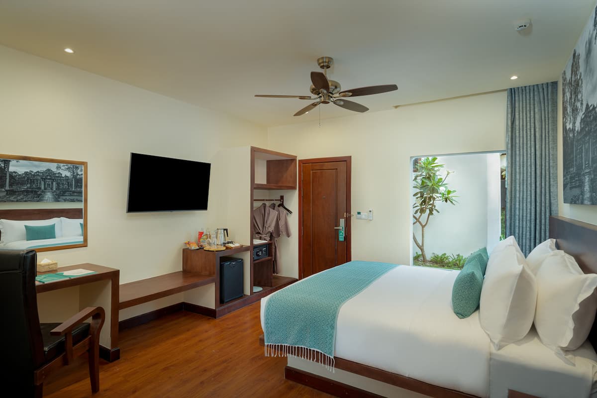 A hotel room with a bed, a desk, a chair, a TV, a ceiling fan, a wardrobe, and a window with a plant.