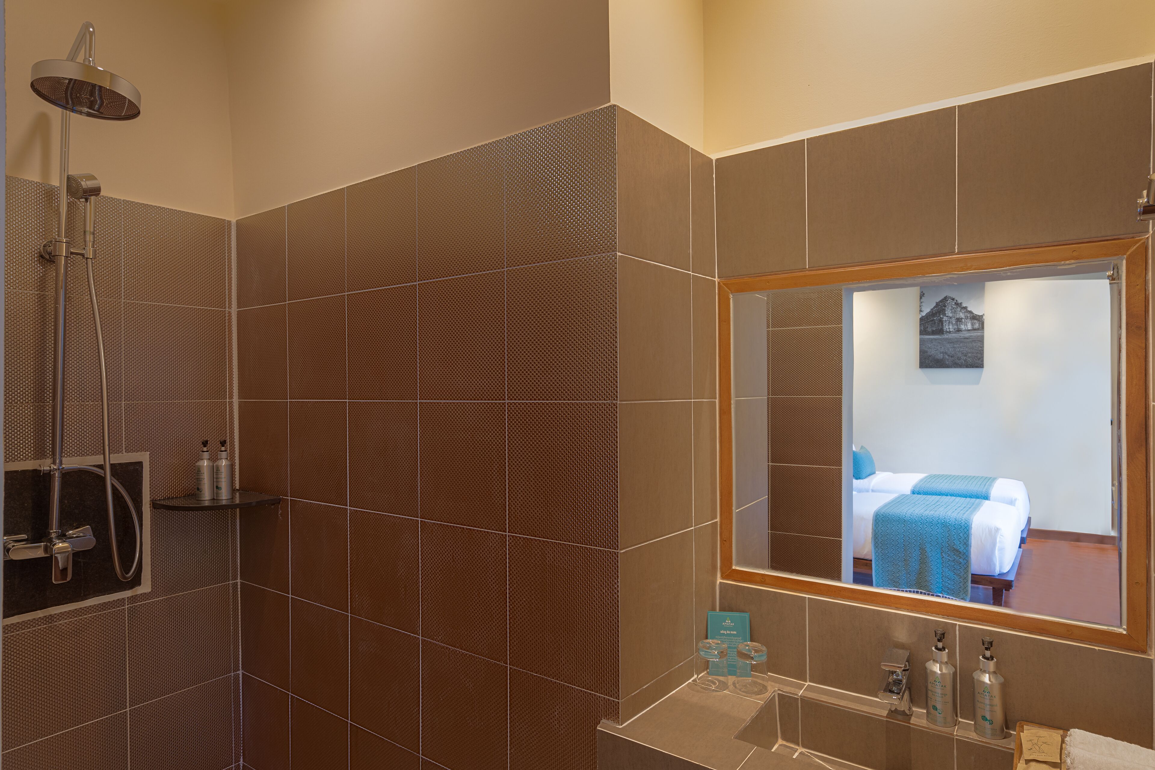 A bathroom with a shower, a sink, and a mirror reflecting a bedroom with a bed and a window.