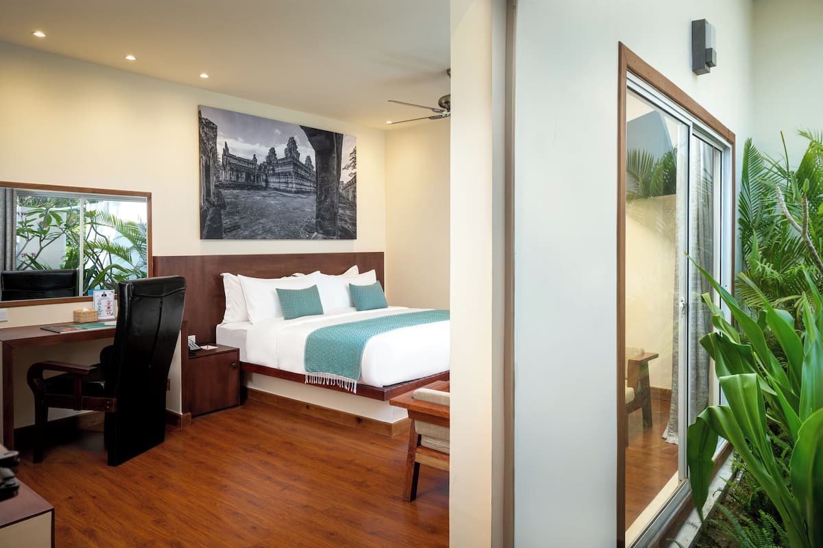 Une chambre d’hôtel moderne avec un lit en bois, un bureau, une chaise, une grande fenêtre donnant sur un paysage verdoyant et un téléviseur fixé au mur.