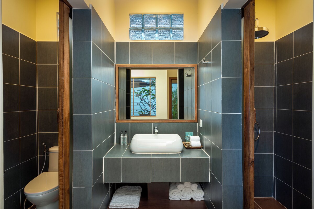 A modern bathroom with dark tiles, a wooden-framed mirror, a white sink, and a toilet.