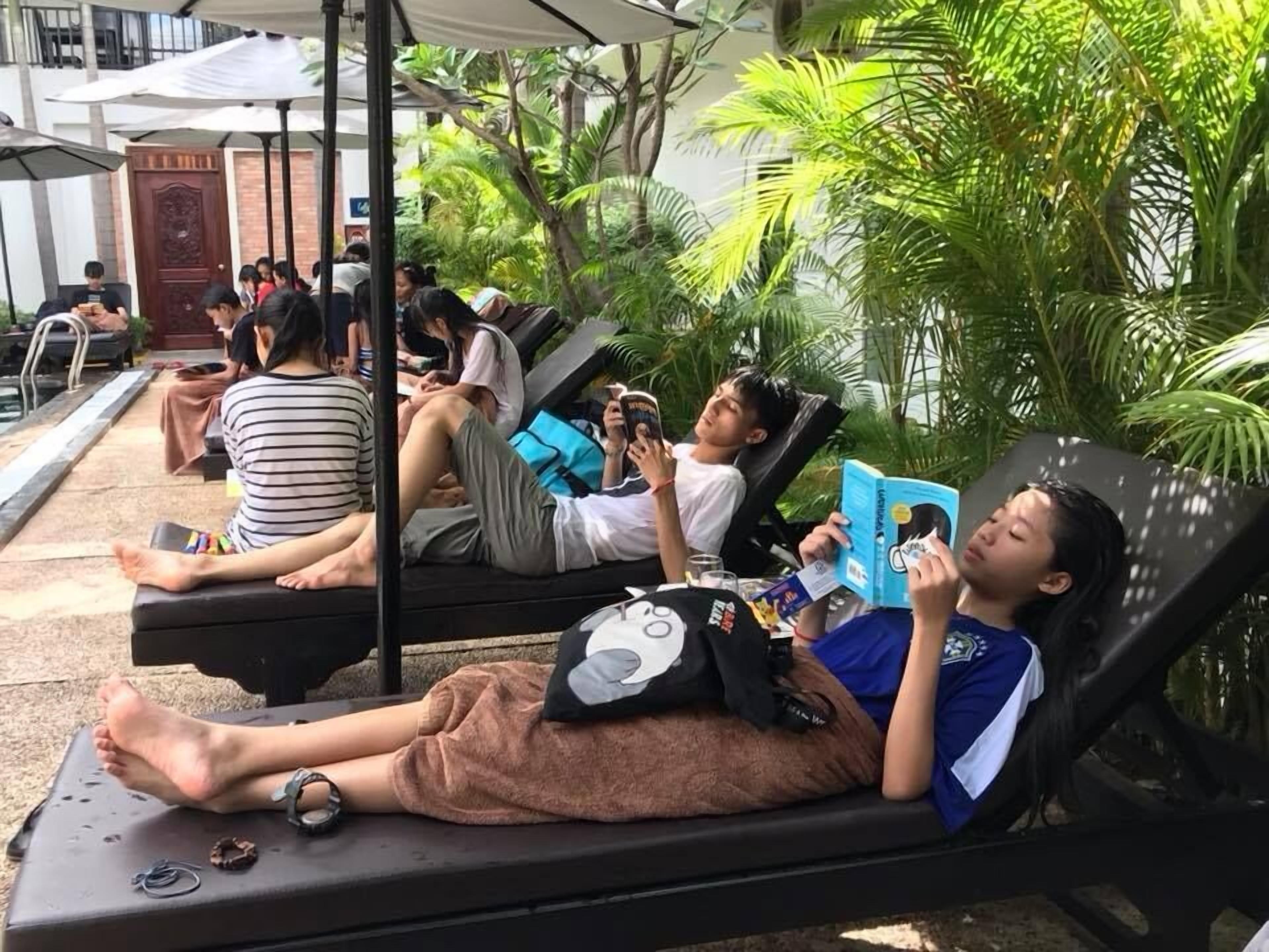 A poolside lounge area with reclining chairs, some occupied by people reading or using their phones, surrounded by lush greenery.