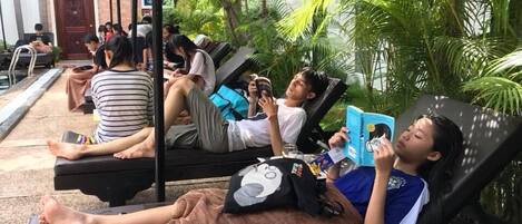 A poolside lounge area with reclining chairs, some occupied by people reading or using their phones, surrounded by lush greenery.