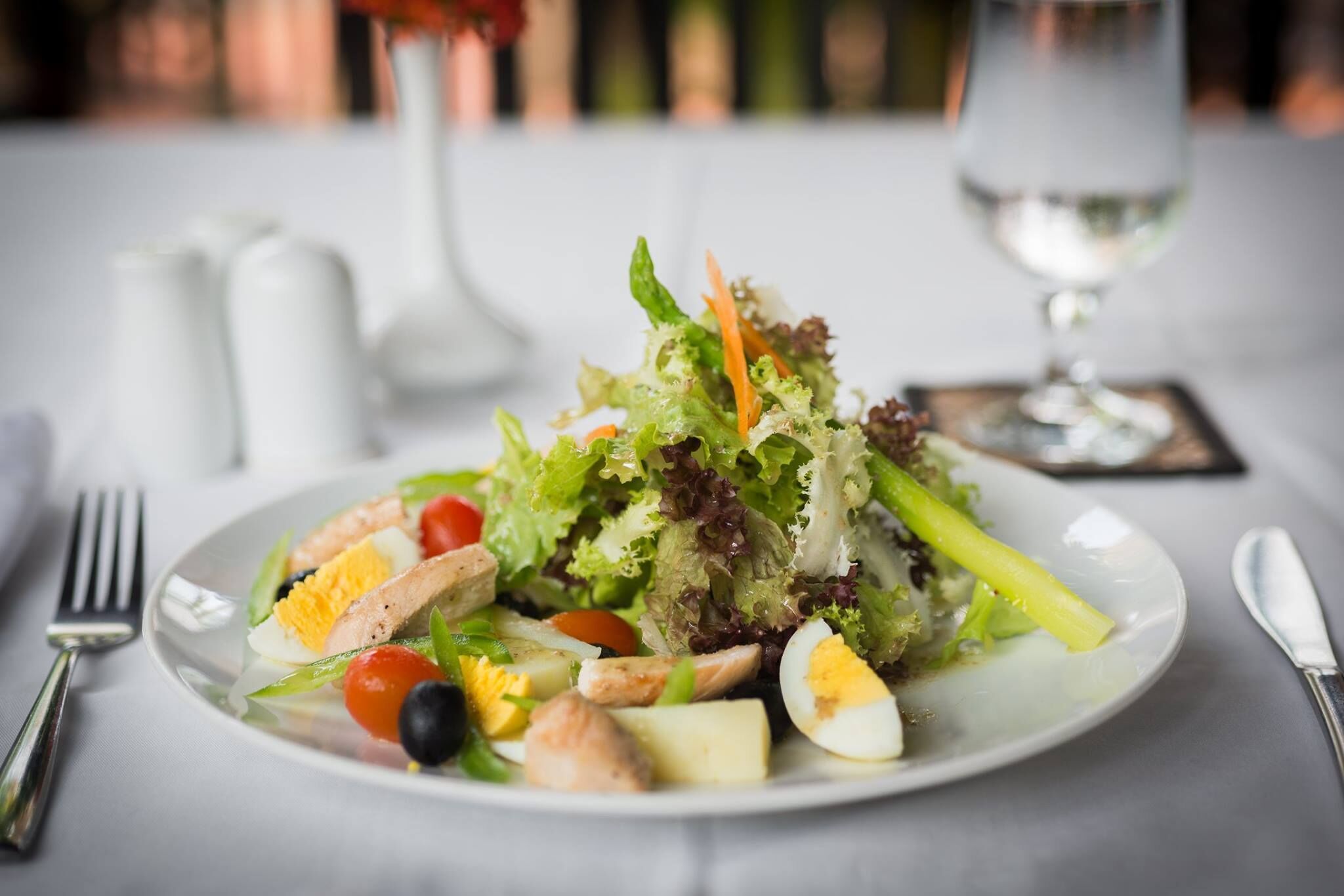 A plate of salad with various vegetables and proteins, accompanied by a glass of water and cutlery on a table.