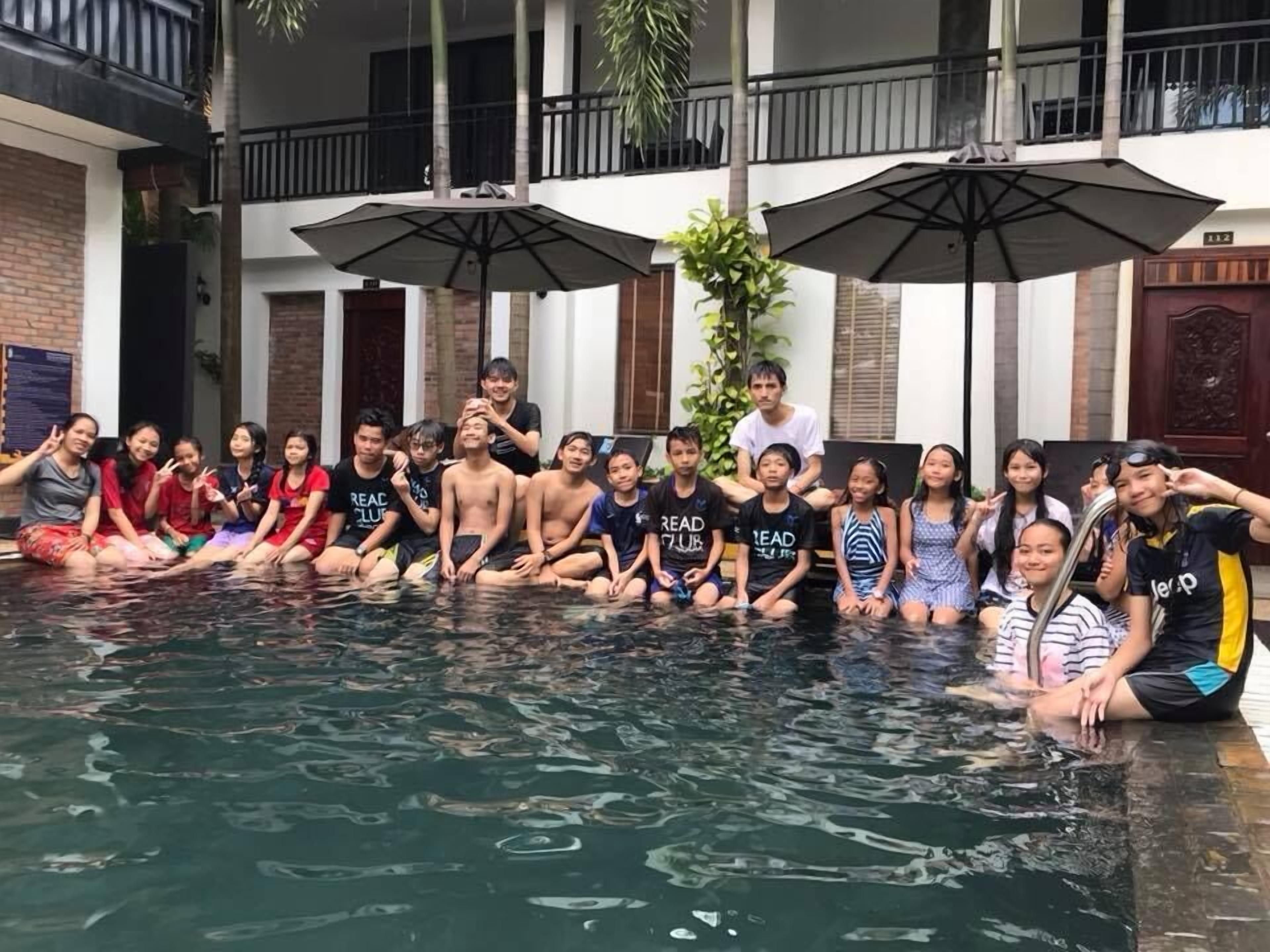 A group of people sitting around a pool with umbrellas and a building in the background.