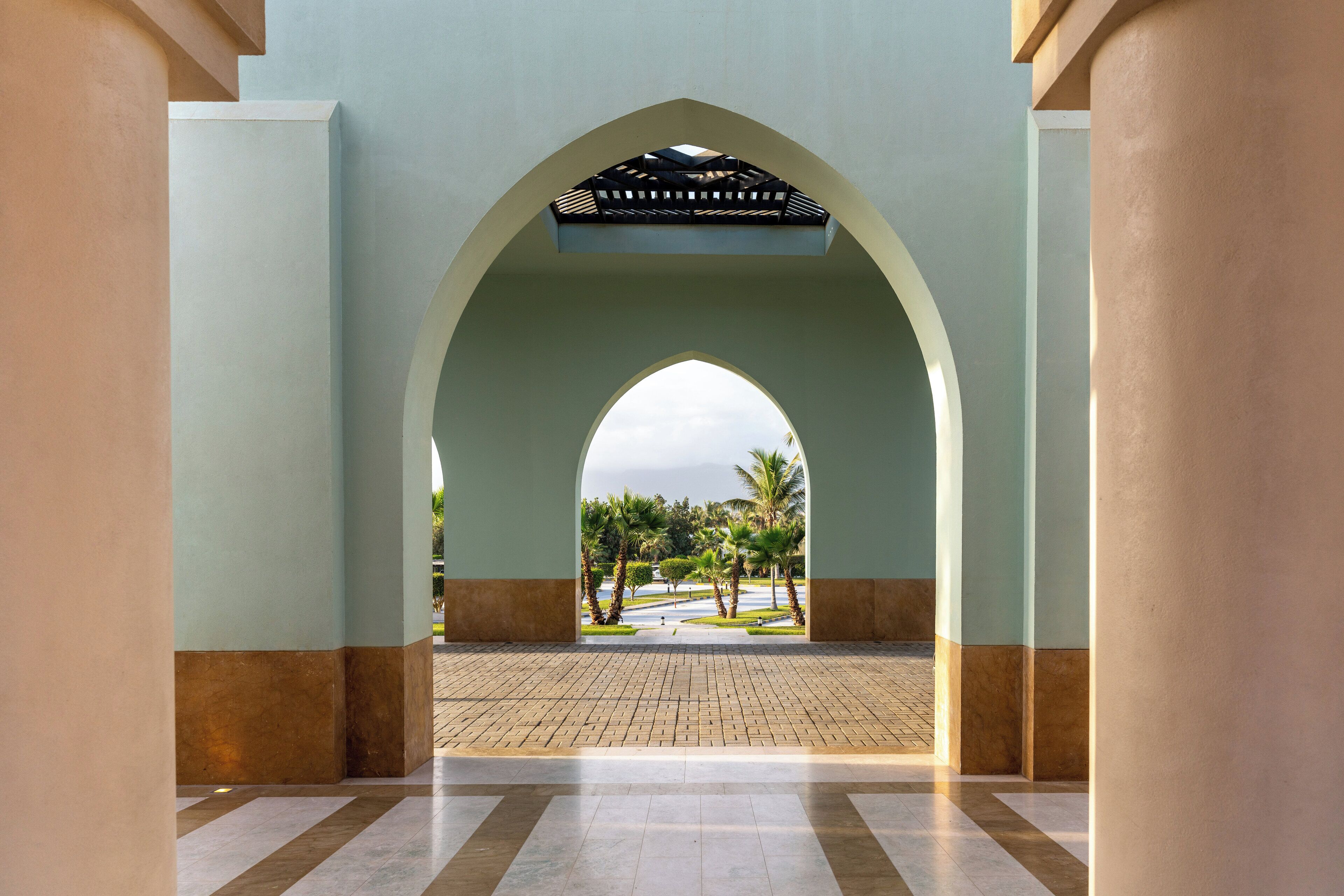 An arched corridor with a tiled floor and a view of palm trees and a pathway outside.
