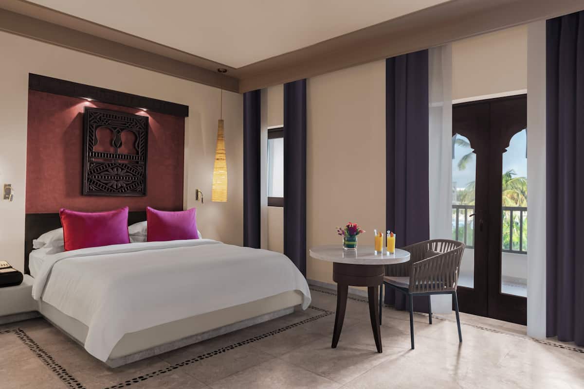 A hotel room with a large bed, a small table with a chair, a decorative wall piece, and a view of the outdoors.