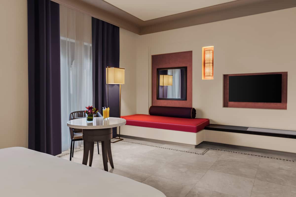 A hotel room with a round dining table, a red bench, a large mirror, and a flat-screen TV mounted on the wall.