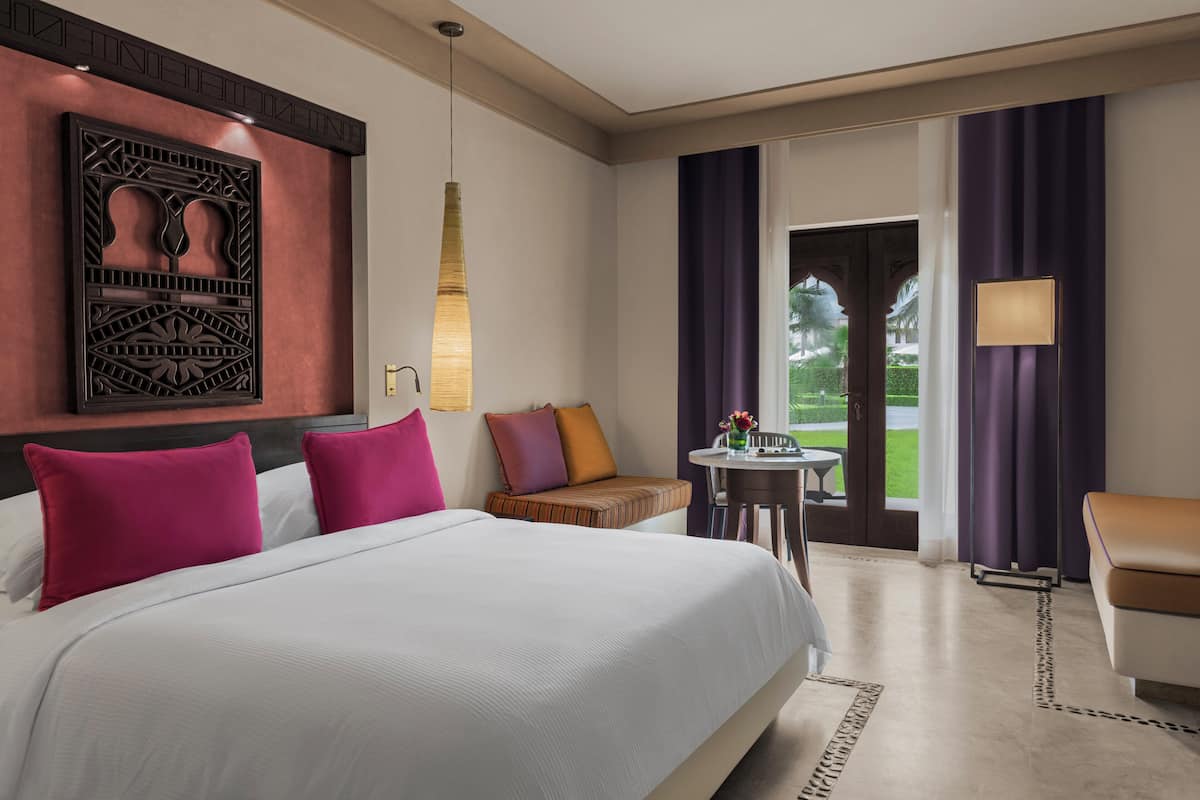 A hotel room with a large bed, a seating area with cushions, a small table with a vase, and a view of the outdoors through a door.