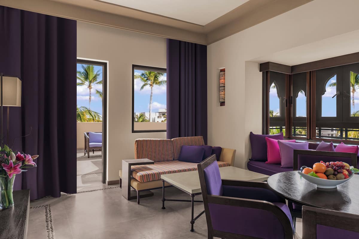 A modern hotel room with a sofa, dining table, and a view of palm trees.