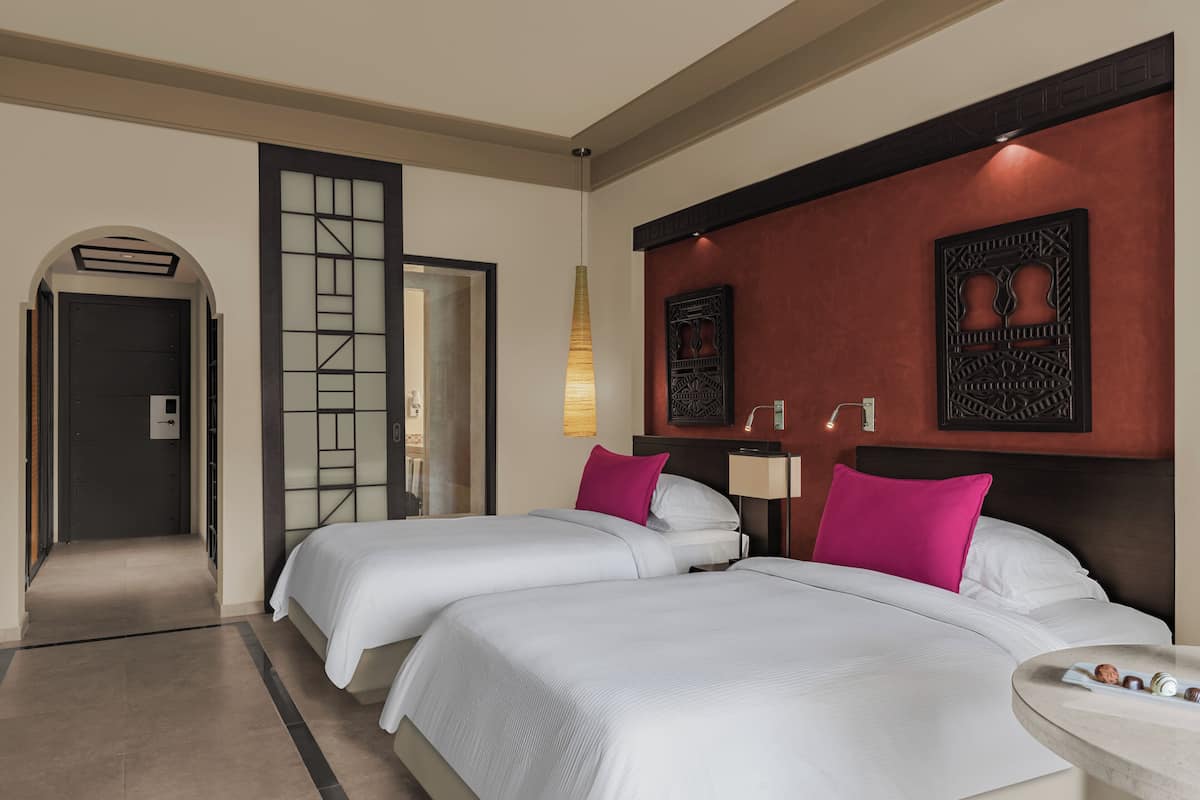 A hotel room with two beds, a small table with snacks, a decorative wall piece, and a door leading to another room.