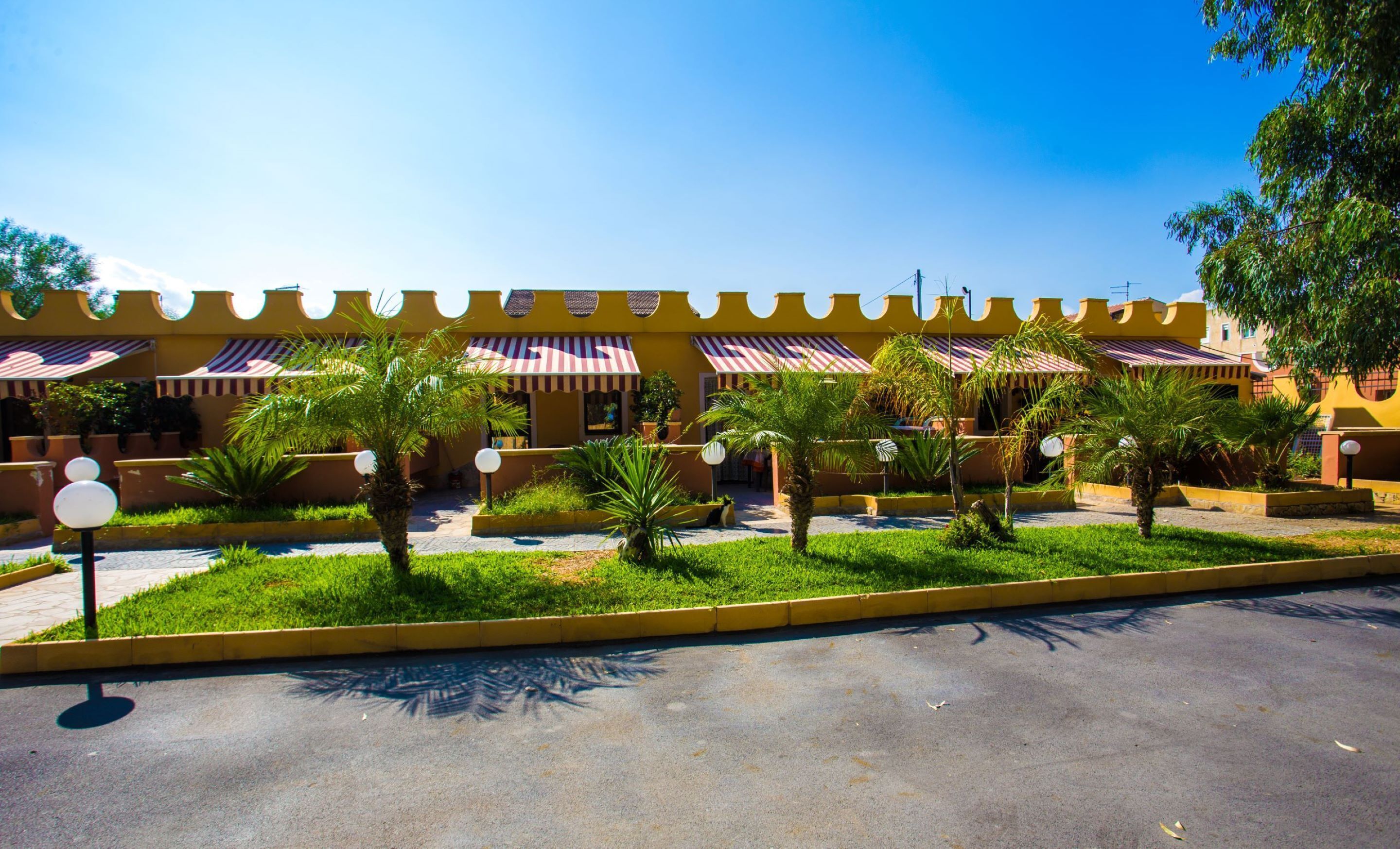 A building with a yellow facade and striped awnings, surrounded by greenery and palm trees.