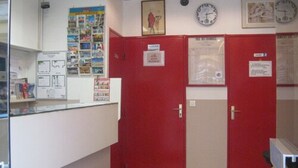 Una zona de recepción con mostrador de vidrio, puertas rojas y beiges, un reloj y un cartel en la pared.