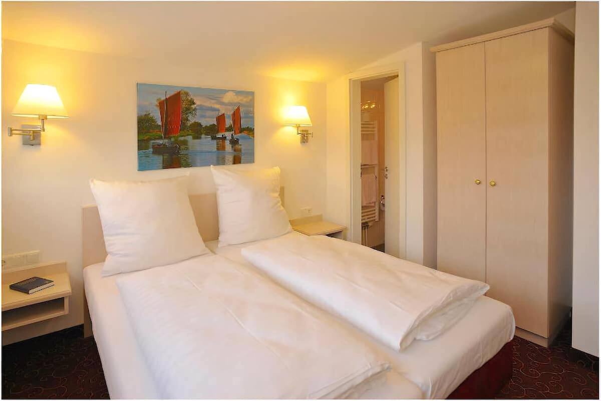 A hotel room with a double bed, two bedside lamps, a painting of a sailboat, and a wardrobe.