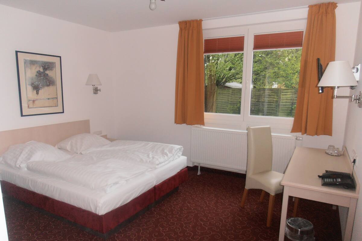A hotel room with a double bed, a desk with a chair, a radiator, and a window with curtains.