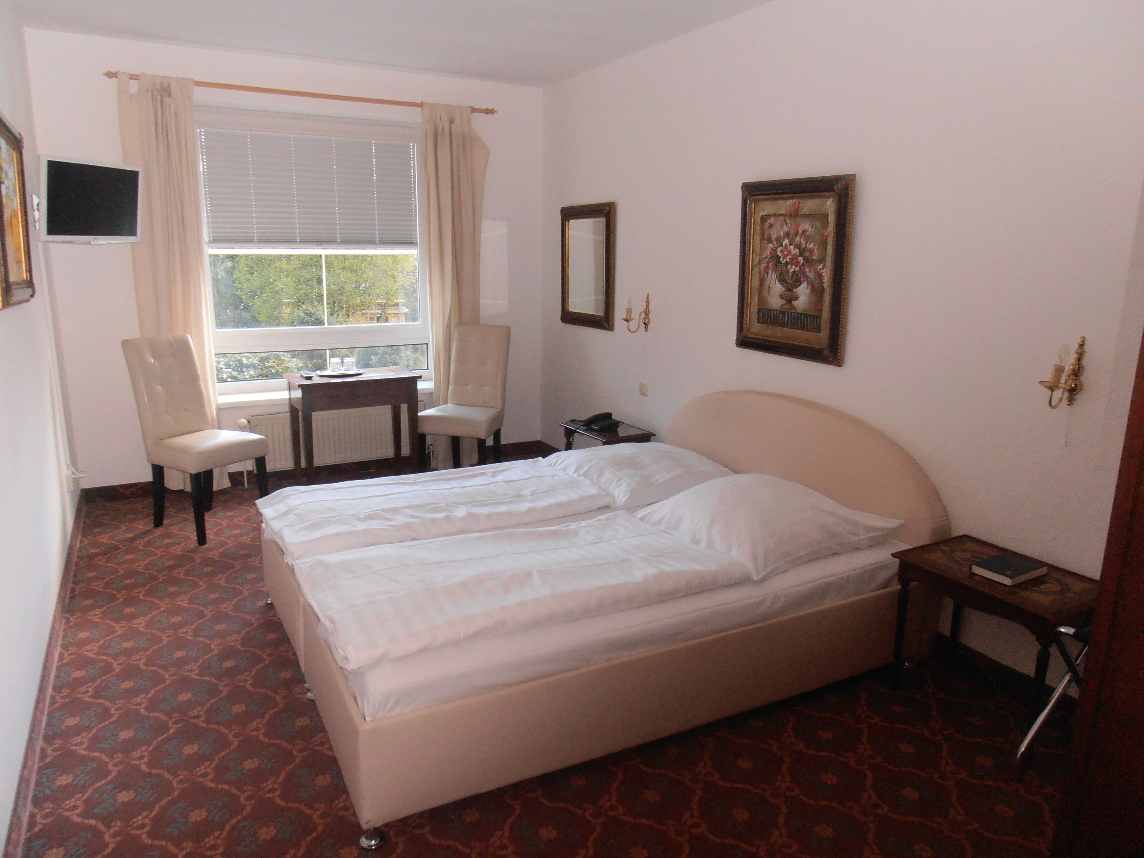 A hotel room with a bed, two chairs, a small table, a window with curtains, and a painting on the wall.