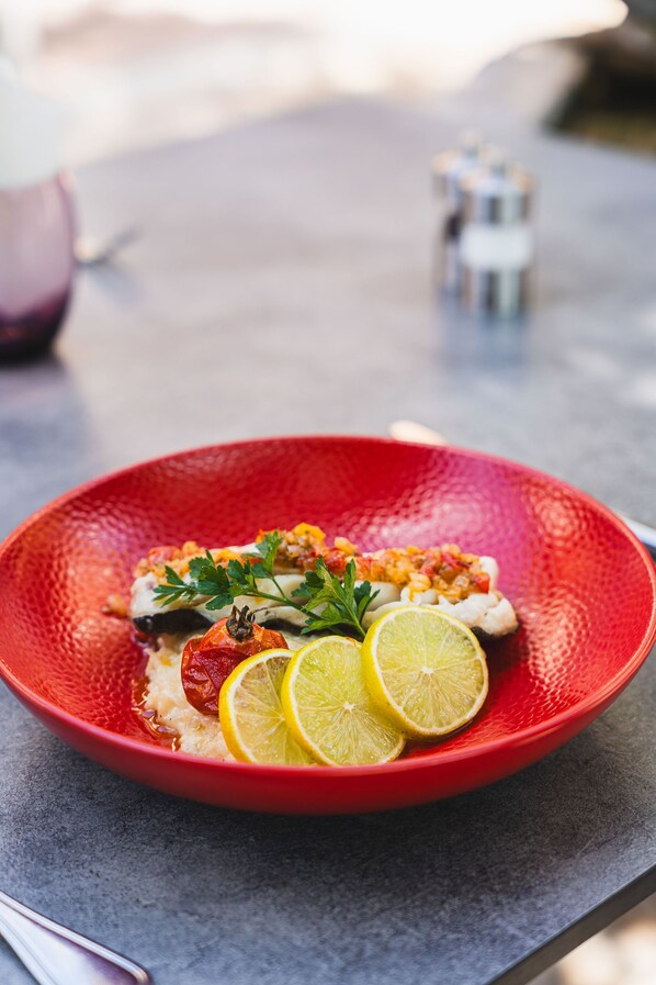 A red textured plate with a white fish dish, garnished with parsley and lemon slices, on a gray surface.