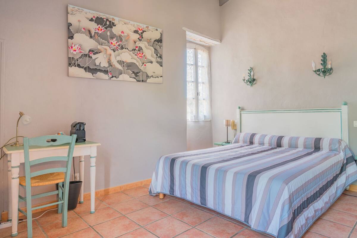 Un dormitorio con una cama, un escritorio y una silla. Hay una ventana con cortinas, un cuadro en la pared y una lámpara de pared.