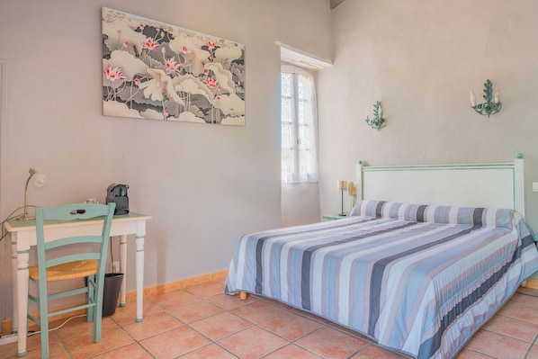 A bedroom with a bed, a desk, and a chair. There is a window with curtains, a painting on the wall, and a wall-mounted lamp.