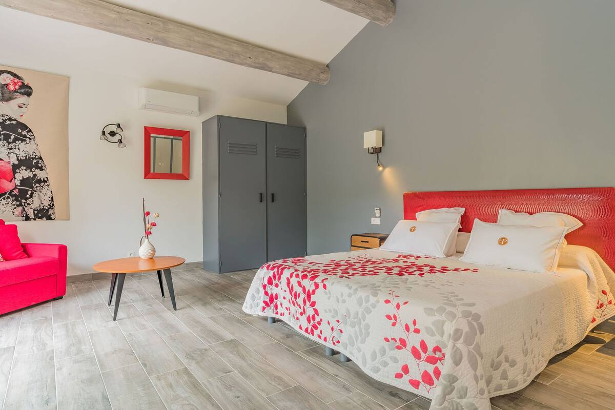 Un dormitorio con cama, cabecera roja, armario gris, silla roja y una mesita de noche con un florero.
