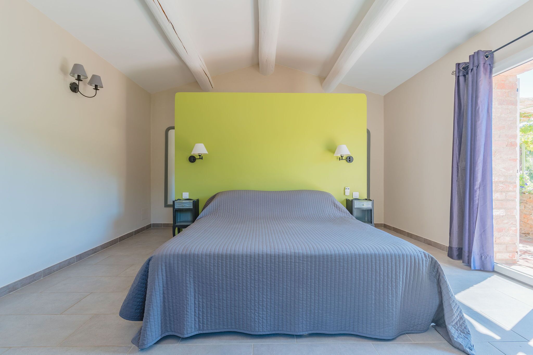 Une chambre à coucher avec un lit, deux lampes de chevet, un mur d’accent vert et une fenêtre avec des rideaux.