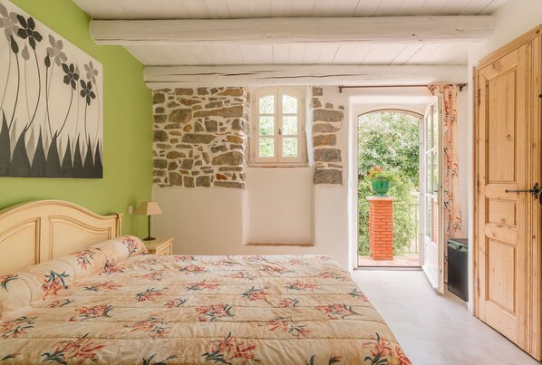 A bedroom with a bed, a nightstand, a painting, a stone wall, a window, and a door leading to a balcony.
