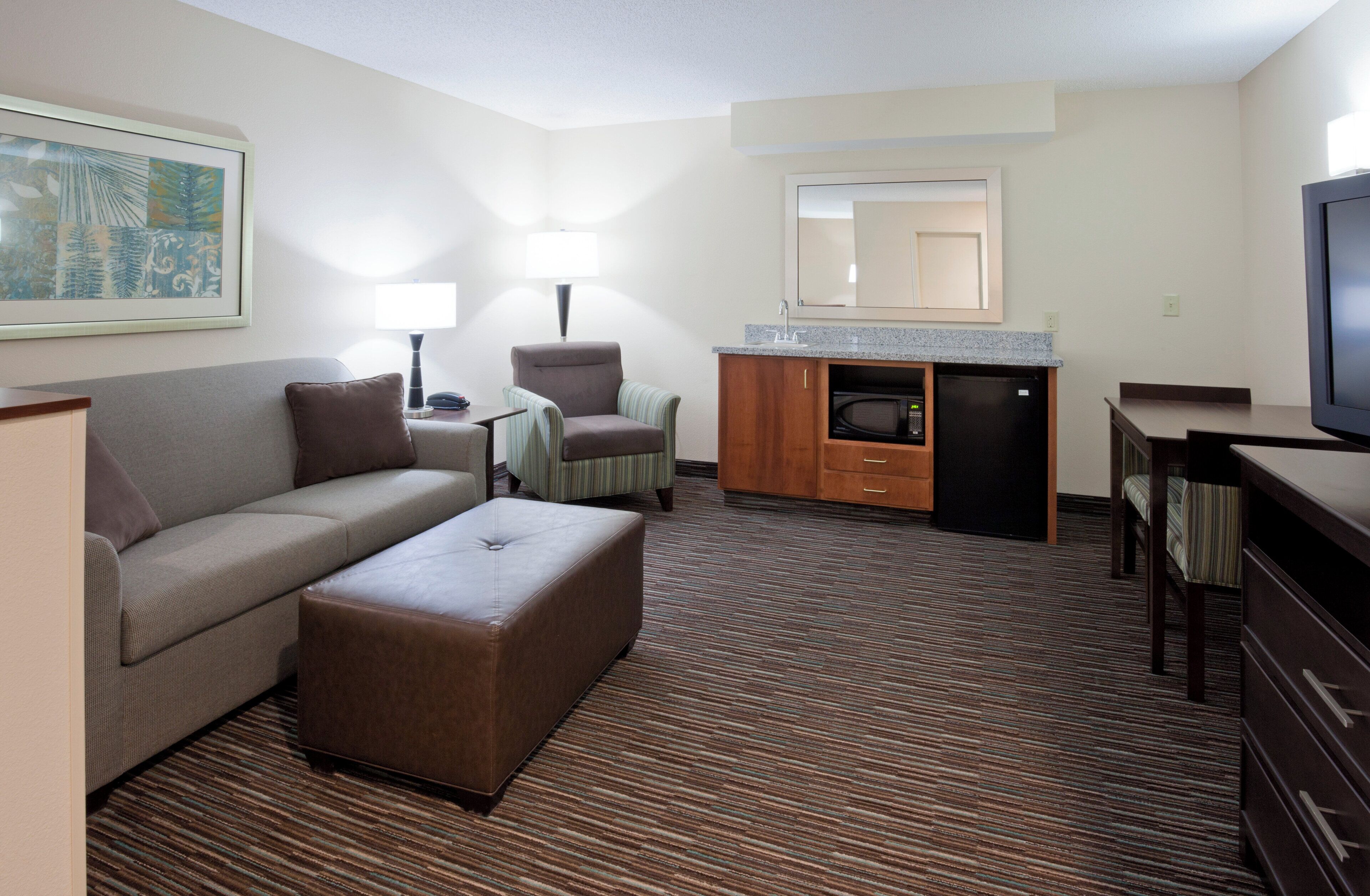 A hotel room with a sofa, armchair, ottoman, kitchenette, and a television.