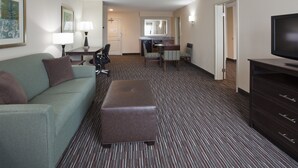 A hotel room with a green sofa, a brown ottoman, a TV, a dining table with chairs, a kitchen area, and a large mirror.