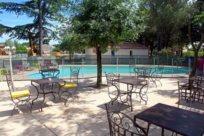 Un área de piscina al aire libre con sillas y mesas de metal, rodeada de árboles y una valla.