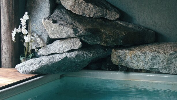 Zona de piscina con borde de madera, una toalla en el borde y rocas grandes al fondo.