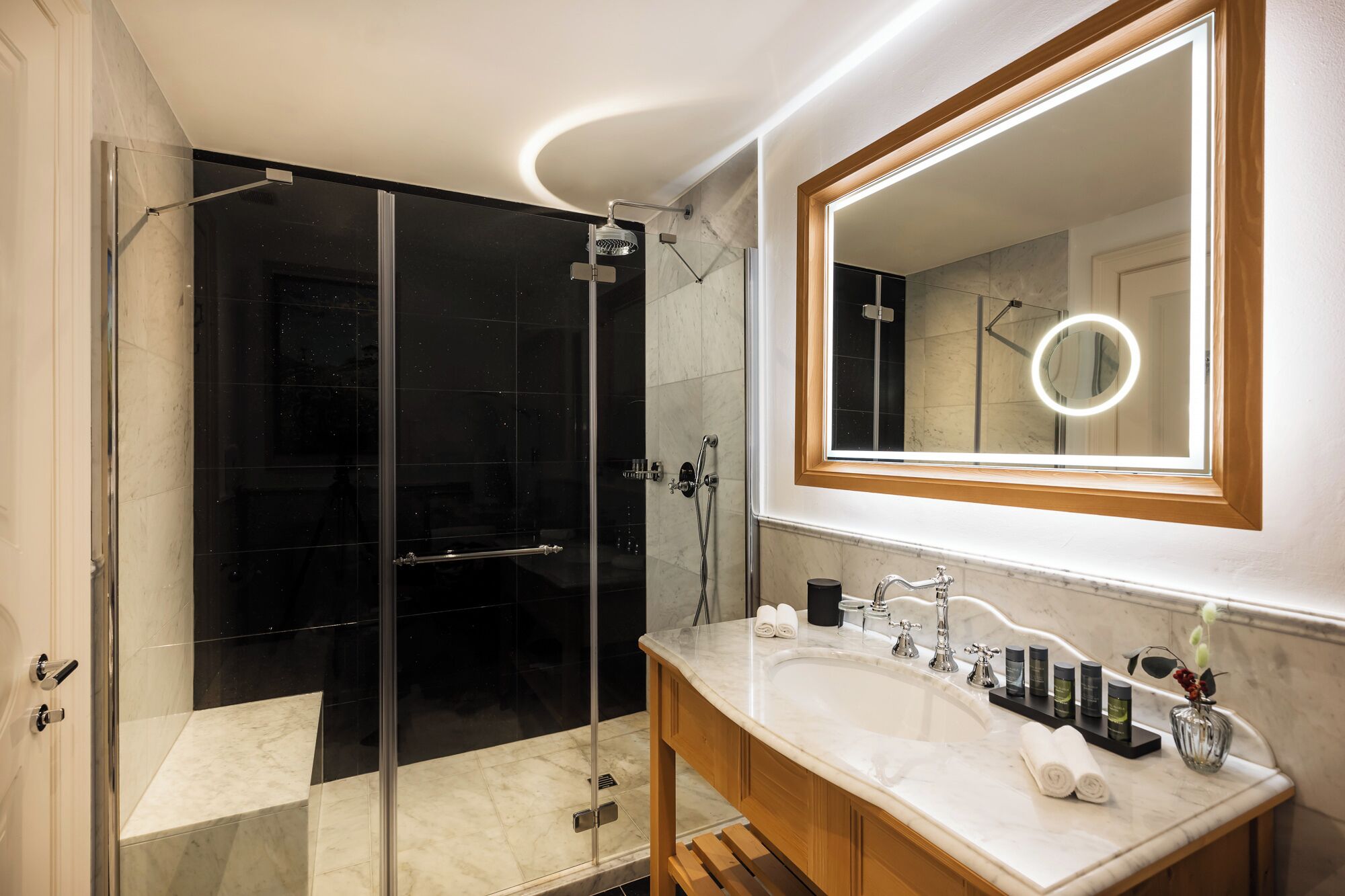 Une salle de bain moderne avec une douche en verre, un grand miroir avec éclairage intégré et un lavabo en marbre agrémenté d’articles de toilette.