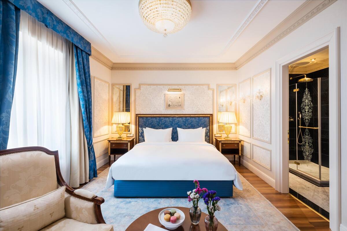 Ett hotellrum med en stor säng, två sängbord med lampor, en stol, ett runt bord med en skål med macarons och en duschhörna.