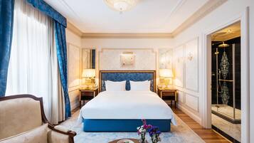 Hotelkamer met een groot bed, twee nachtkastjes met lampen, een stoel, een ronde tafel met een schaal macarons, en een douchehoek.