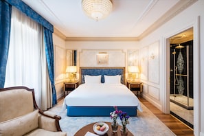 A hotel room with a large bed, two bedside tables with lamps, a chair, a round table with a bowl of macarons, and a shower area.