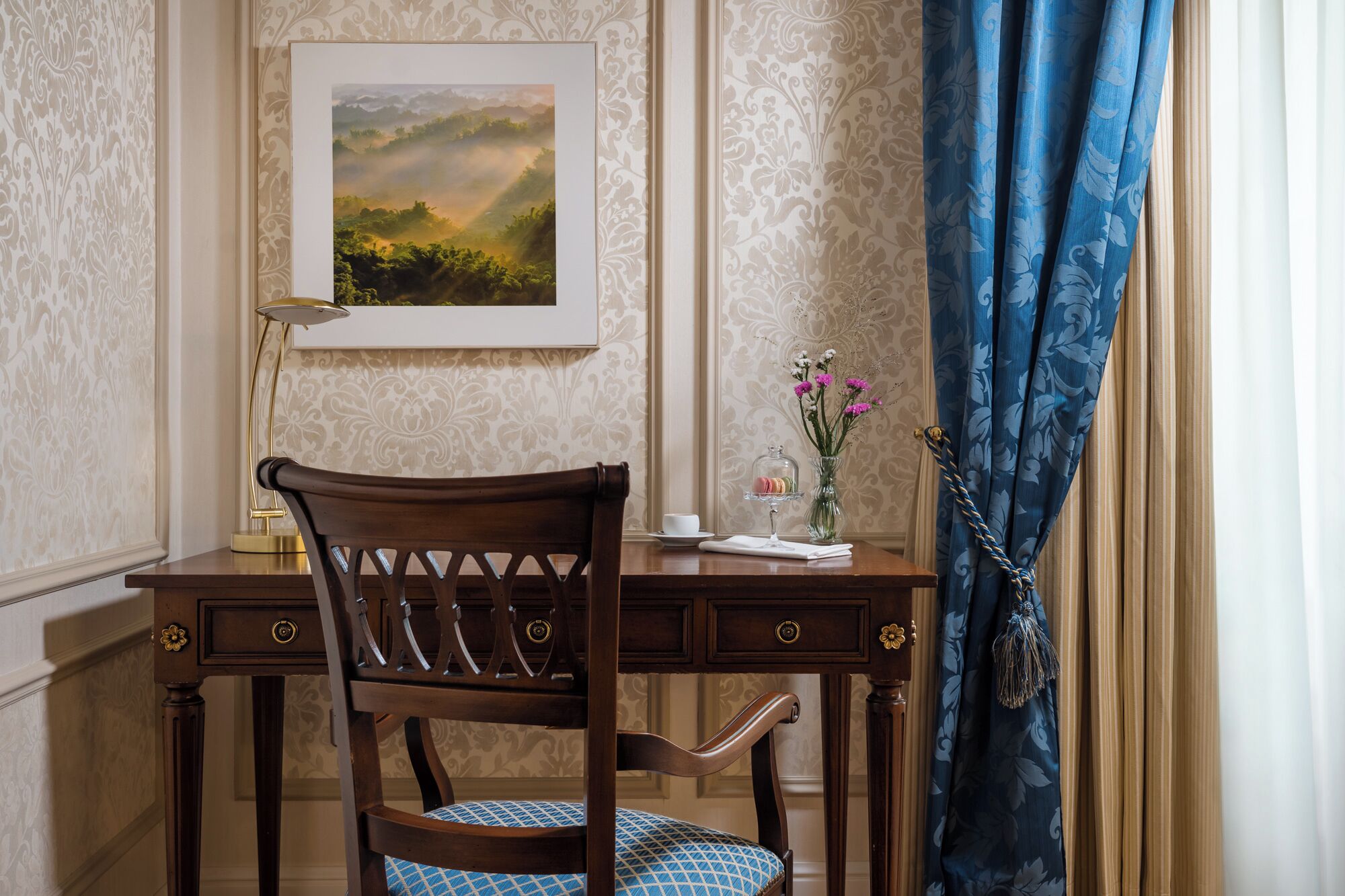 A wooden desk with a chair, a framed landscape painting, a vase with flowers, and a blue curtain.