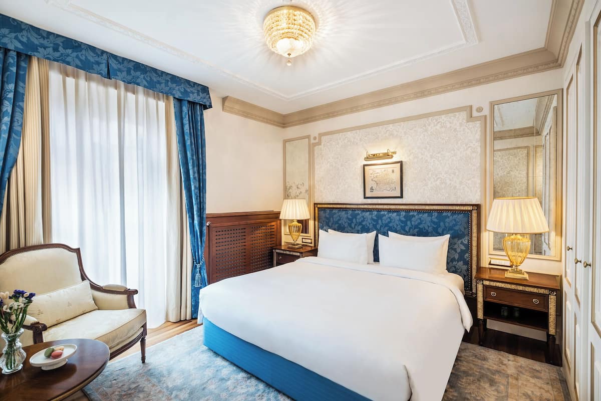 Ett hotellrum med en stor säng, ett träbord vid sängen med lampor, en stol och en vas med blommor.