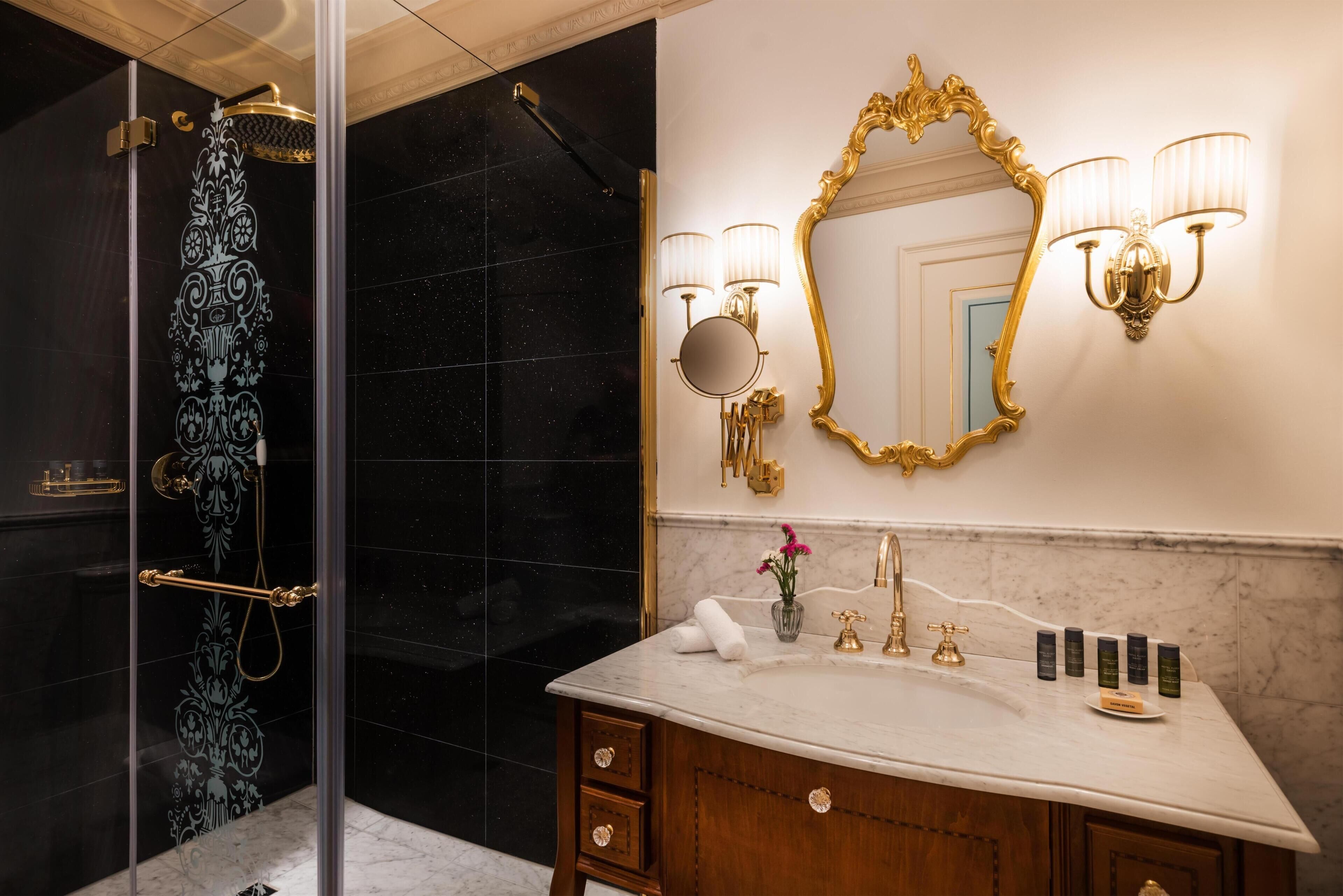 A bathroom with a glass-enclosed shower, a marble sink, a gold-framed mirror, and wall-mounted lighting.