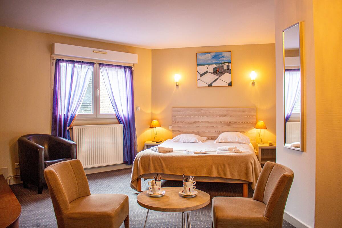 A hotel room with a bed, two chairs, a small table with cups, and a painting on the wall.