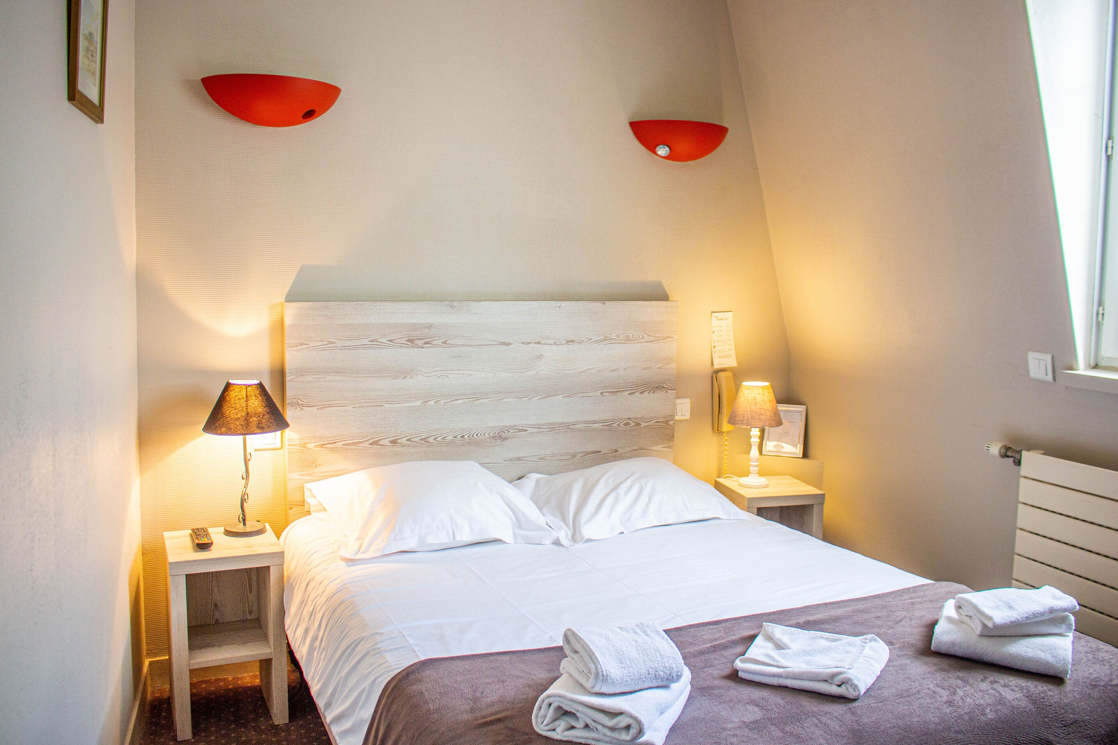A neatly made bed with white linens and a folded towel on the bed. There are two bedside tables, each with a lamp and a phone. The room has a modern design with a wooden headboard and red wall sconces.