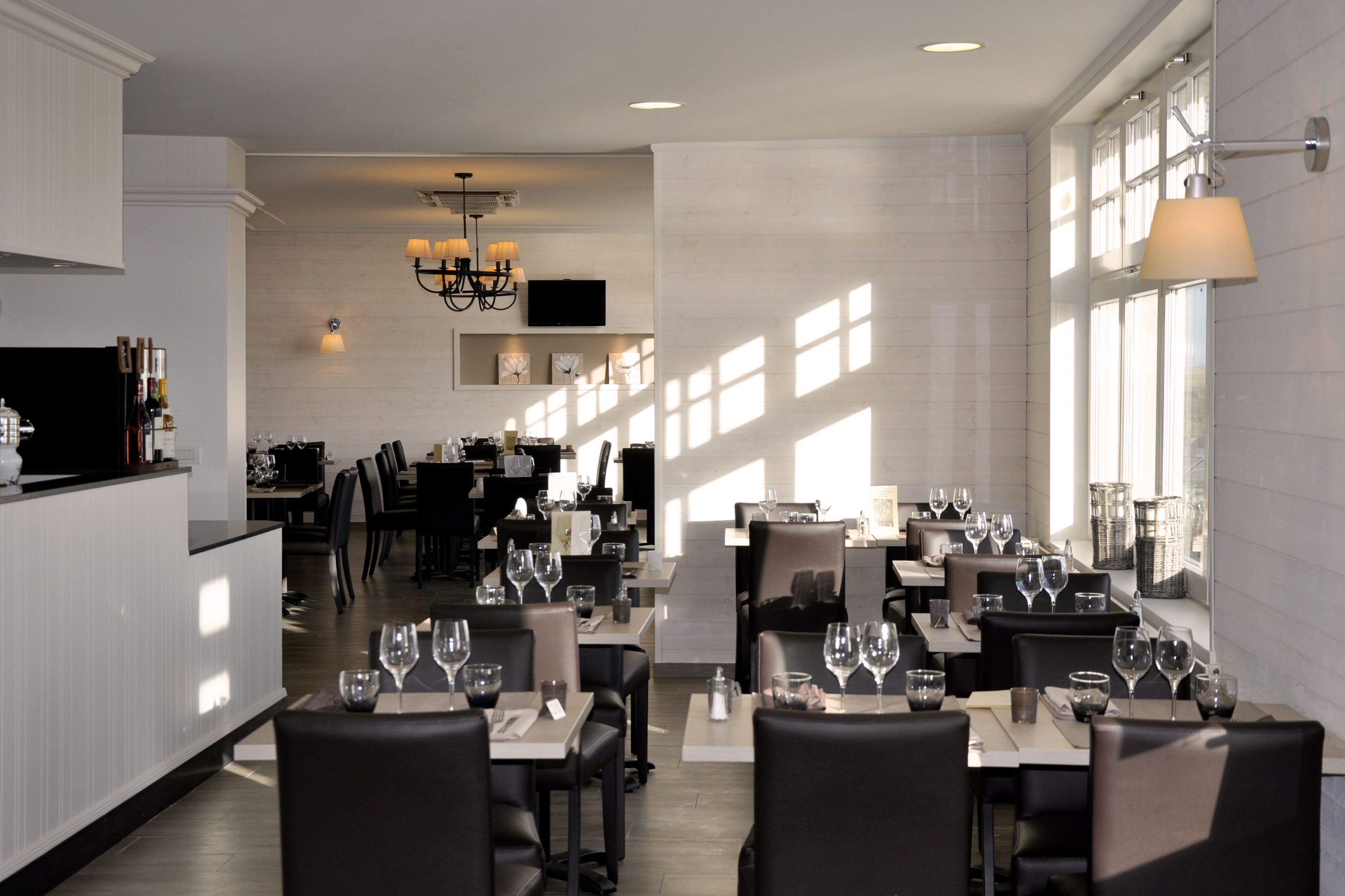 A modern restaurant with a clean, minimalist interior, featuring a long dining area with tables set for multiple guests, a bar area, and a television mounted on the wall.