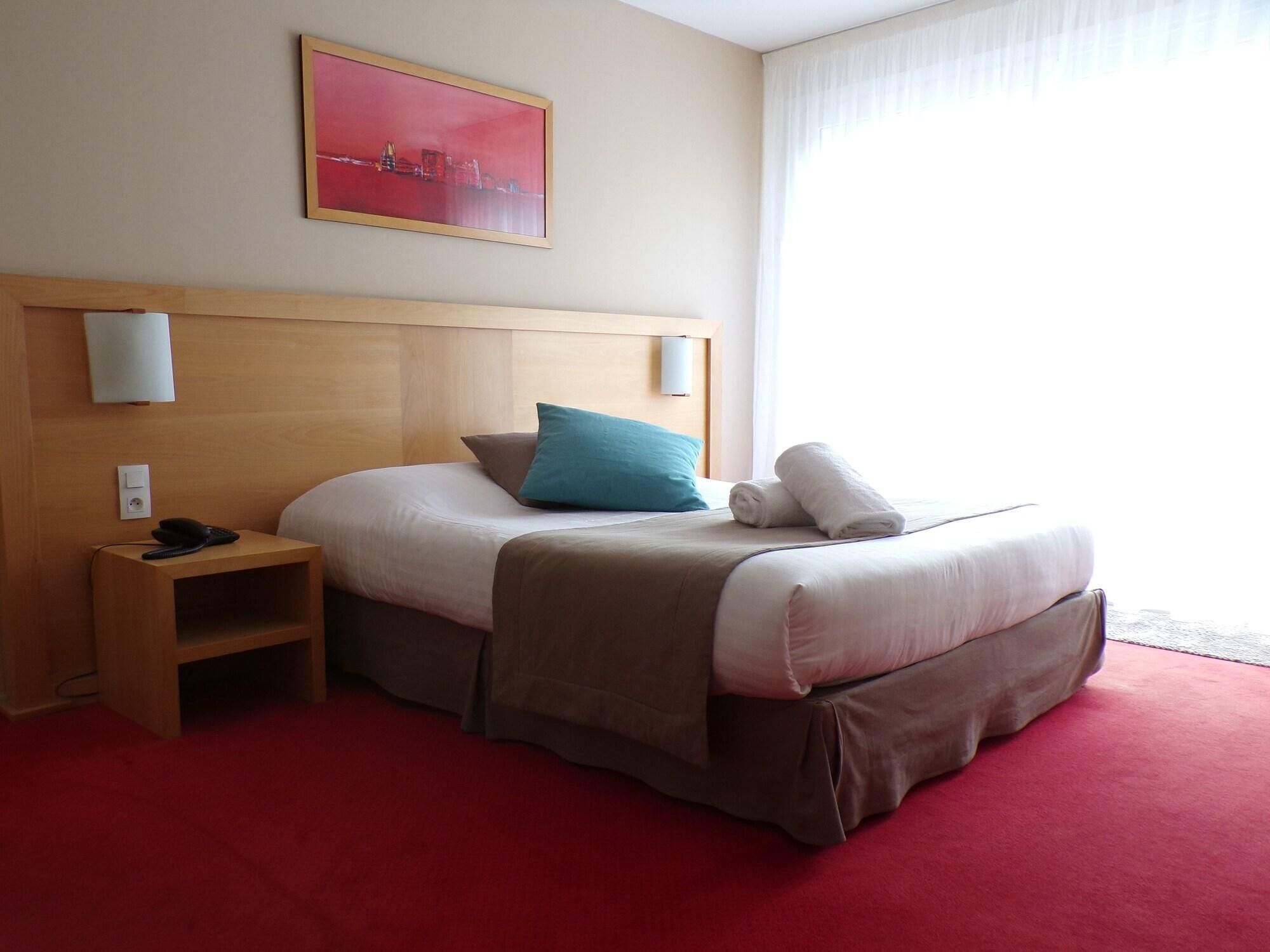 A hotel room with a bed, a nightstand, a lamp, a painting on the wall, and a window with curtains.