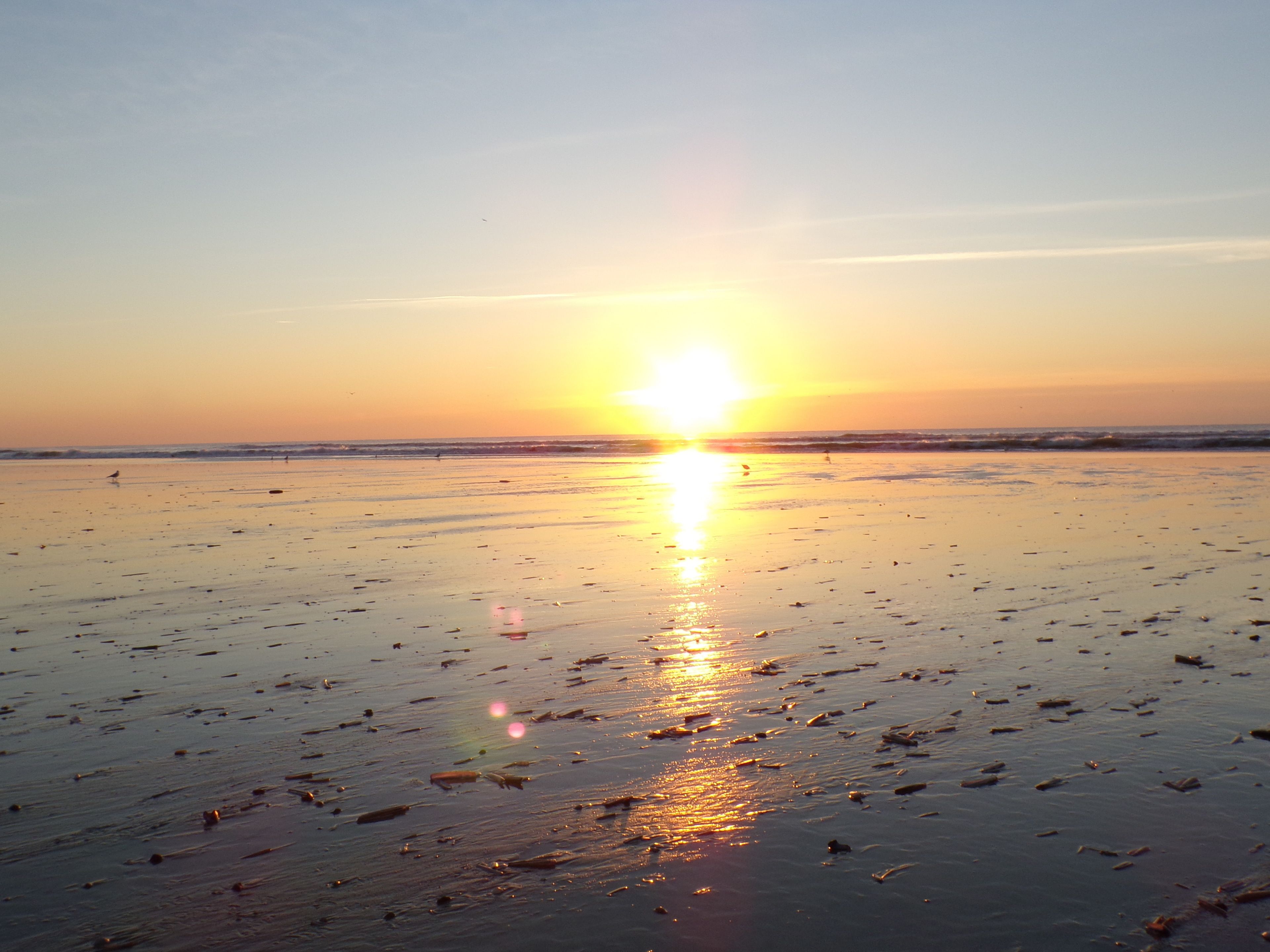 A beach at sunset with the sun low on the horizon, reflecting on the wet sand.