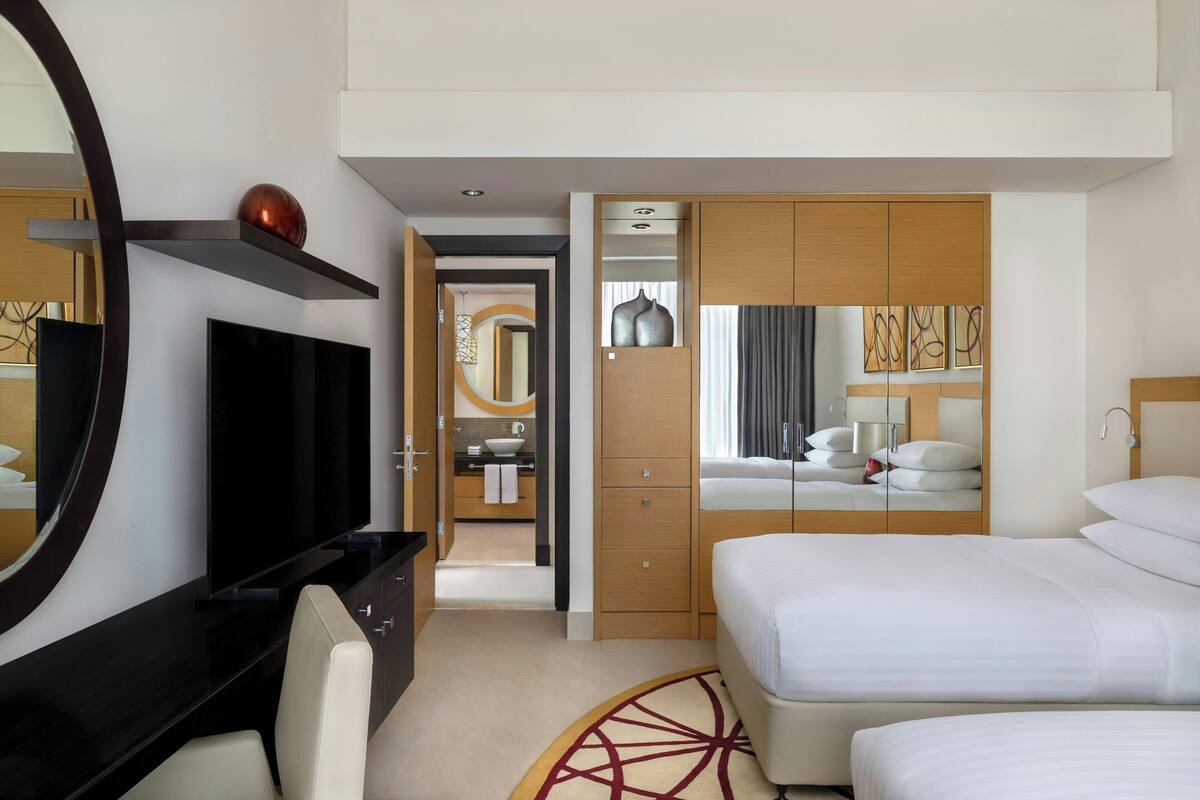 A hotel room with a large mirror, a flat-screen TV, a bed with white bedding, a wooden wardrobe, and a bathroom visible through an open door.
