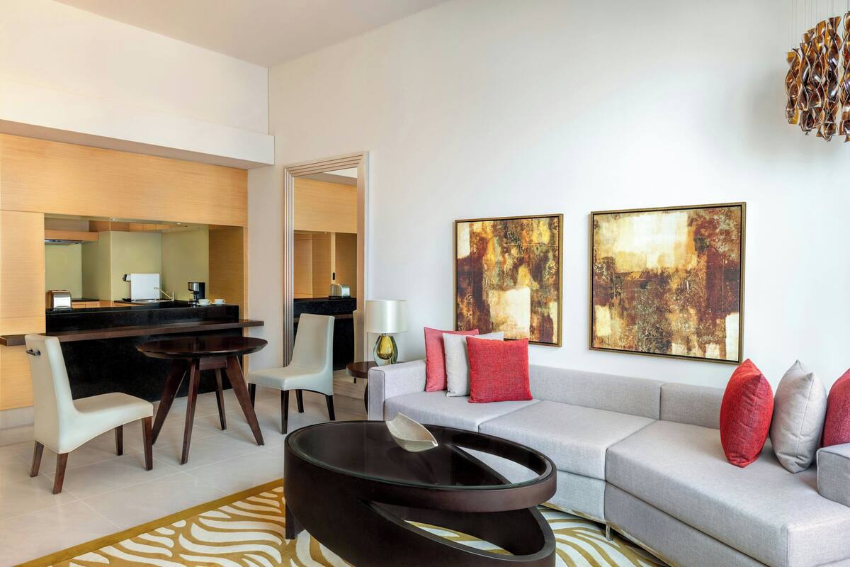 A modern living room with a grey sofa, a round coffee table, and a dining area with a table and chairs. There are abstract paintings on the wall and a unique chandelier.