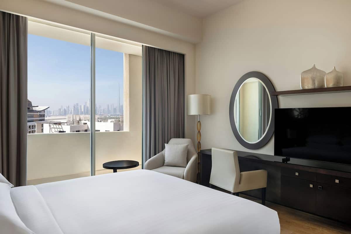 A hotel room with a large bed, a TV, a chair, a small table, and a view of the city skyline.